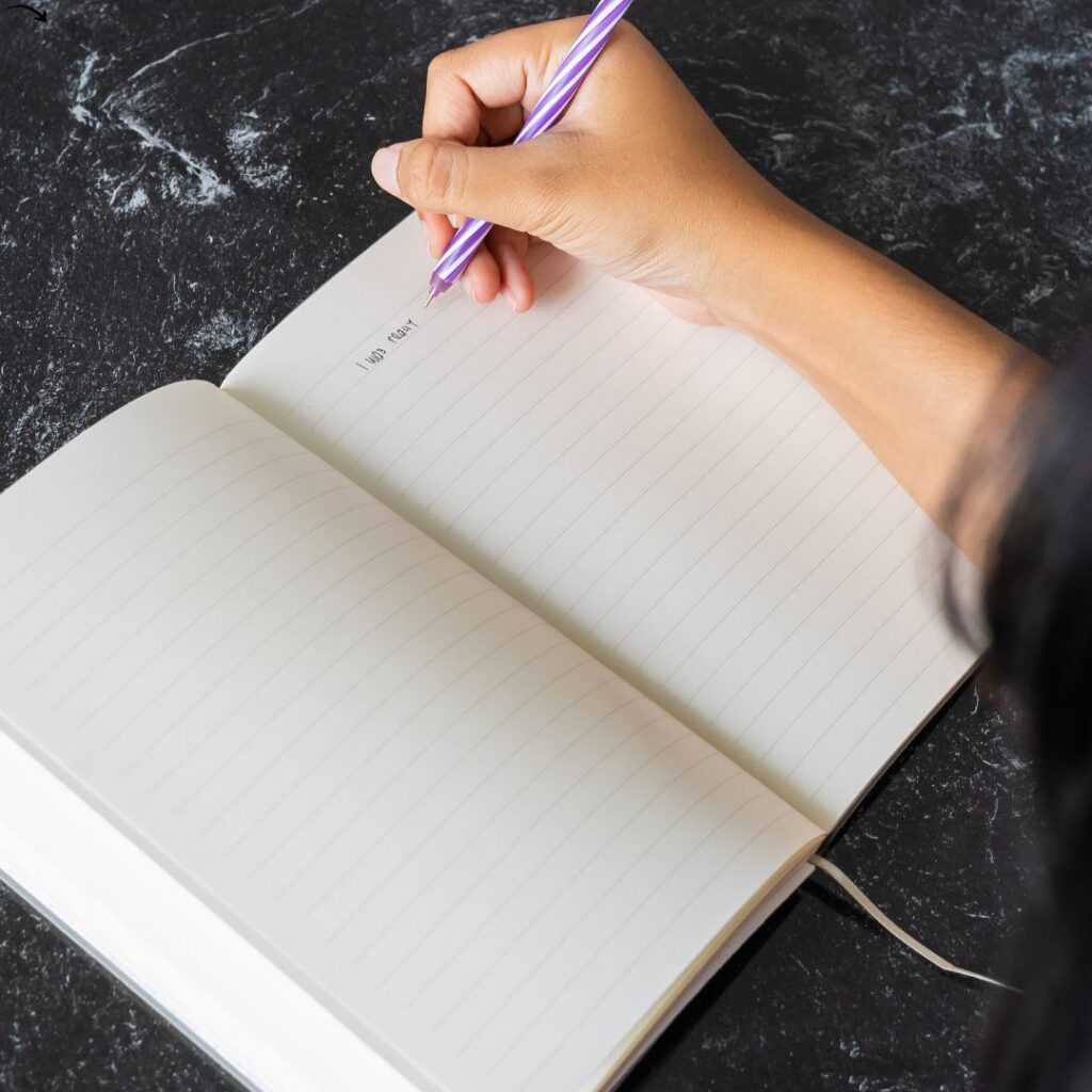 Hand schreibt mit lila Stift in ein Notizbuch auf dunklem Marmor